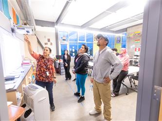 A teacher is pointing to a poster on the wall to the guests