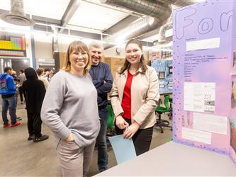A family of 3 are smiling in front of a cardboard display